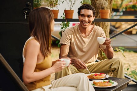Foto de Una pareja interracial disfrutando de una comida juntos en un banco, saboreando su comida en un ambiente acogedor e íntimo. - Imagen libre de derechos