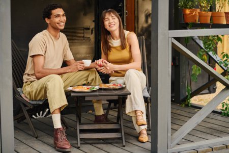 A man and woman of different races are sitting peacefully on a porch, enjoying each others company in a tranquil setting.