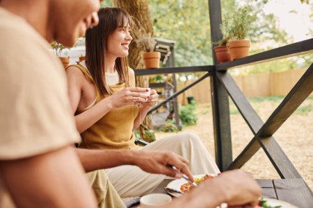 Téléchargez les photos : Un couple interracial profite d'un moment paisible sur un banc, partageant un repas ensemble dans un parc pittoresque. - en image libre de droit