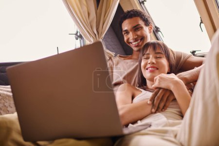 Téléchargez les photos : Couple interracial assis sur le lit, absorbé dans l'écran d'ordinateur portable dans le camping-car moderne - en image libre de droit