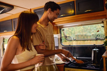 An interracial couple is joyfully preparing a meal together inside a cozy camper van.