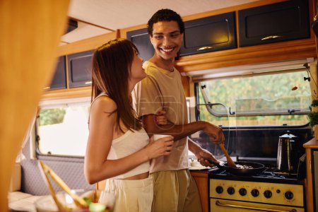 Ein gemischtrassiges Paar kocht zusammen in einem Wohnmobil auf einem romantischen Wochenende, umgeben von gemütlicher Atmosphäre.
