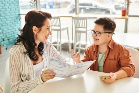 Photo for Good looking happy woman in casual clothes with her son with Down syndrome holding menu and phone - Royalty Free Image