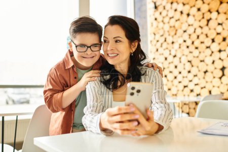 Téléchargez les photos : Gai garçon avec trisomie 21 passer du temps avec sa belle mère dans un café, tenant smartphone - en image libre de droit