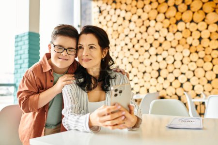 Téléchargez les photos : Joyeux garçon avec trisomie 21 passer du temps avec sa belle mère dans un café, tenant smartphone - en image libre de droit