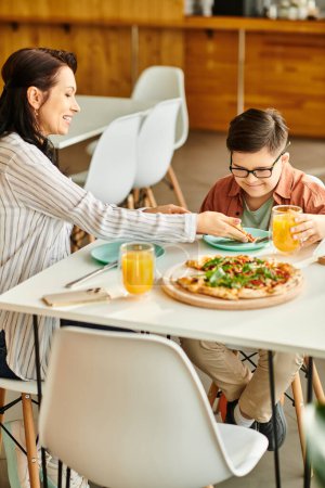 Foto de Atractiva madre comiendo pizza y bebiendo jugo con su hijo lindo inclusivo con síndrome de Down - Imagen libre de derechos