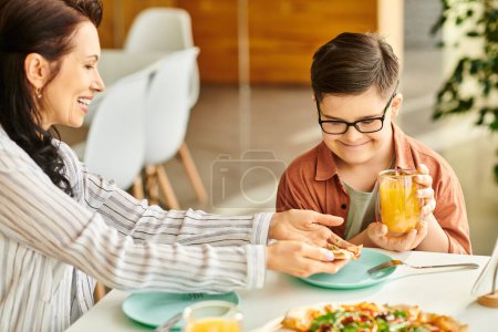 Téléchargez les photos : Belle mère manger de la pizza et boire du jus avec son fils mignon inclusif avec trisomie 21 - en image libre de droit
