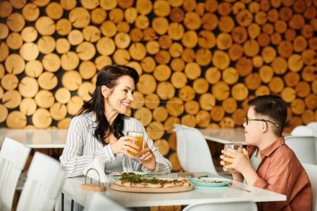 Téléchargez les photos : Mère heureuse manger de la pizza et boire du jus avec son fils mignon inclusif avec trisomie 21 - en image libre de droit