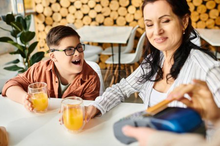 schöne fröhliche Mutter beim Bezahlen mit Kreditkarte neben ihrem inklusiven Sohn mit Down-Syndrom im Café