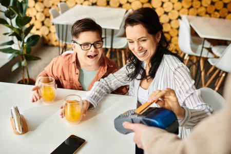 schöne fröhliche Mutter beim Bezahlen mit Kreditkarte neben ihrem inklusiven Sohn mit Down-Syndrom im Café