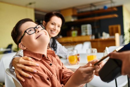 entzückender inklusiver Junge mit Down-Syndrom bezahlt mit Smartphone im Café in der Nähe seiner fröhlichen Mutter