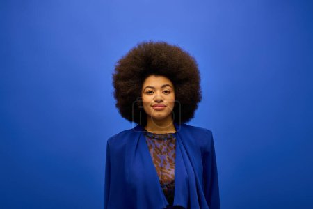 Foto de Elegante mujer afroamericana con el pelo rizado de pie con confianza sobre un fondo azul vibrante. - Imagen libre de derechos