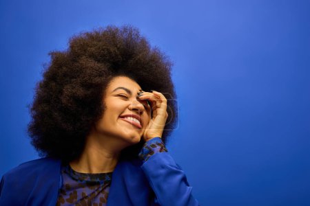 Stilvolle Afroamerikanerin mit lockigem Haar lächelt und berührt ihr Gesicht vor einer lebendigen Kulisse.
