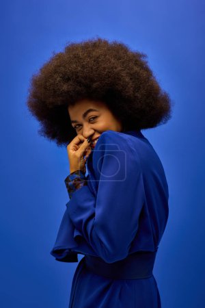 A fashionable African American woman with curly hairdoposes in front of a bright blue backdrop.