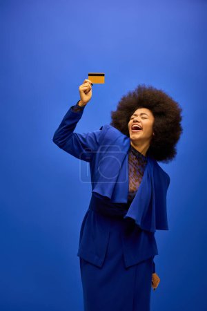 Stylish African American woman holding credit card in blue suit against vibrant backdrop.