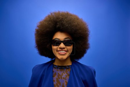 Téléchargez les photos : Femme afro-américaine élégante pose dans une veste bleue à la mode et des lunettes de soleil sur fond vibrant. - en image libre de droit