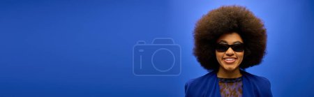 Téléchargez les photos : Femme afro-américaine à la mode avec des cheveux bouclés et des lunettes de soleil, portant une veste bleue, frappant une pose. - en image libre de droit