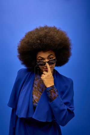 Stylish African American woman posing in trendy blue coat and glasses on vibrant backdrop.