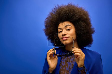 Téléchargez les photos : Une femme afro-américaine élégante tenant des lunettes de soleil jusqu'à son visage dans un contexte vibrant. - en image libre de droit