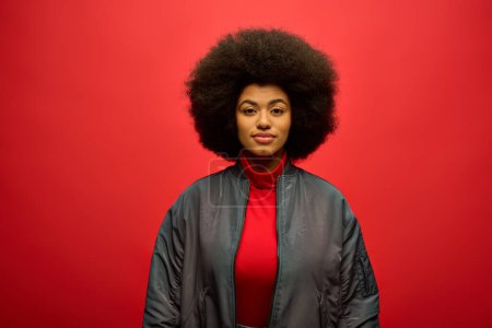Stylish African American woman with curly hairdostands confidently against bright red backdrop.