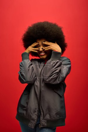 Stylish African American woman in trendy attire covering face with hands.