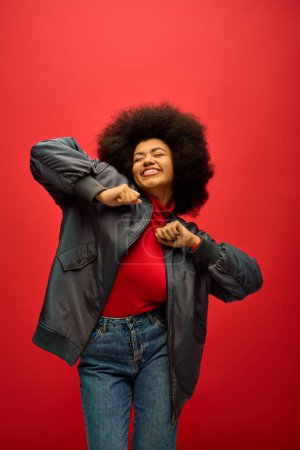 Stilvolle Afroamerikanerin mit lockigem Haar steht selbstbewusst vor leuchtend rotem Hintergrund.