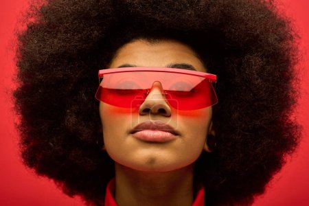 Téléchargez les photos : Une femme afro-américaine élégante avec des lunettes de soleil rouges bouclées pose sur une toile de fond vibrante. - en image libre de droit