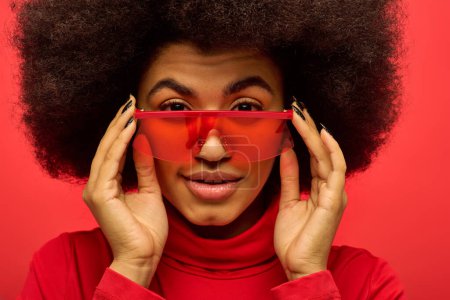 A stylish African American woman with curly hairdohairstyle holds up a pair of red glasses.
