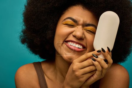 Téléchargez les photos : Une femme afro-américaine élégante tient le pinceau sur son visage dans un contexte vibrant. - en image libre de droit