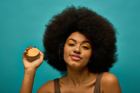 Stylish African American woman with curly hairdoholding cream on vibrant backdrop.