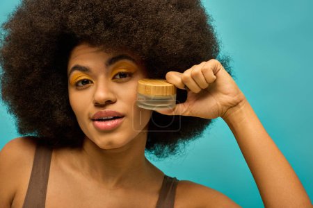 Elegante mujer afroamericana con el pelo rizado posando con un frasco de maquillaje.