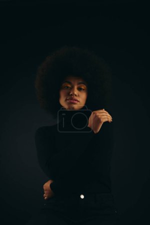 A fashionable African American woman with curly hairdostands confidently in the dark.
