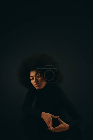 Femme afro-américaine élégante assise dans une pièce sombre.