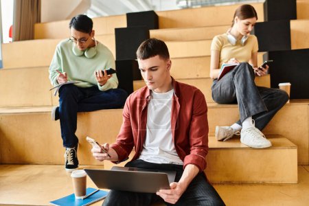 A man, surrounded by a multicultural group of students, sits on steps, engrossed in a laptop, absorbed in his digital studies.