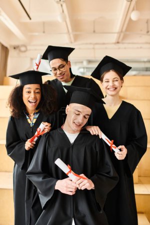 Groupe diversifié d'étudiants en robes de remise des diplômes et casquettes académiques posant heureusement pour une image à l'intérieur.
