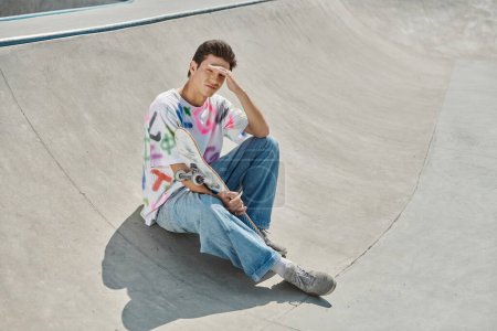 Ein junger Skater-Junge sitzt an einem sonnigen Tag friedlich auf seinem Skateboard in einem lebhaften Skatepark.