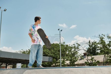 Ein junger Skaterboy hält ein Skateboard in der Hand, während er an einem sonnigen Sommertag selbstbewusst auf einer Rampe in einem lebhaften Skatepark steht.