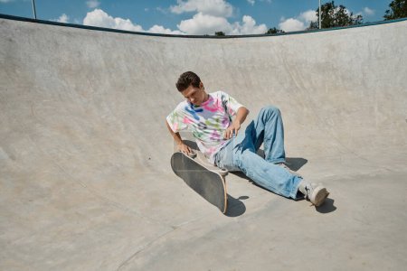 Foto de Un joven patinador desafía la gravedad, sentado cerca del monopatín en una rampa en un parque de skate al aire libre en un día soleado de verano.. - Imagen libre de derechos
