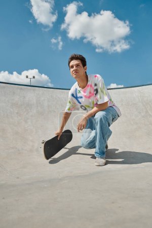Un joven se arrodilla con un monopatín en la mano en un soleado parque de skate.