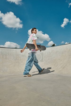 A young man casually walks with a skateboard in hand, exuding a cool and carefree vibe in a summer skate park setting.