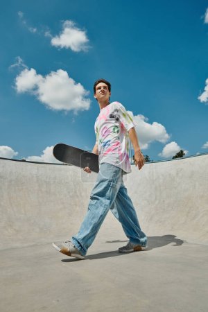 Téléchargez les photos : Un jeune homme marchant à travers un skate park, skateboard à la main, profitant de la journée d'été. - en image libre de droit
