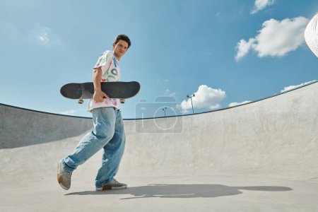 A young man confidently walking in a skate park, holding his skateboard under the bright sun.