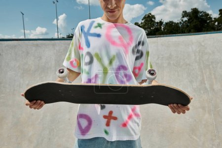 Foto de Un joven sostiene con confianza un monopatín en sus manos en un parque de skate en un día soleado de verano. - Imagen libre de derechos