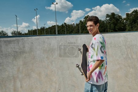 A young man confidently holds his skateboard in a vibrant skate park on a sunny summer day.