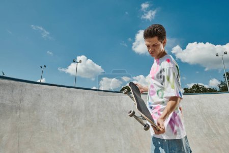 Foto de Un joven sostiene un monopatín en un parque de skate en un día soleado, mostrando su pasión por el monopatín. - Imagen libre de derechos