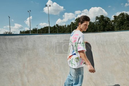 A young skater boy holds a skateboard at a skate park on a sunny summer day.