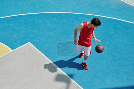 Ein junger Mann steht auf einem Basketballfeld und hält einen Ball in der Hand, um an einem sonnigen Tag zu spielen.
