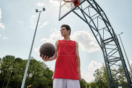 Foto de Un joven con una camisa roja se levanta al aire libre, sosteniendo una pelota de baloncesto en su mano en un día soleado de verano.. - Imagen libre de derechos