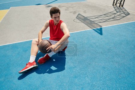 Ein junger Mann sitzt an einem sonnigen Sommertag gedankenverloren auf einem Basketballplatz und hält einen Basketball.