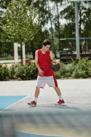 Foto de Un joven se para en una cancha de baloncesto, sosteniendo una pelota en su mano, listo para jugar bajo el sol de verano. - Imagen libre de derechos
